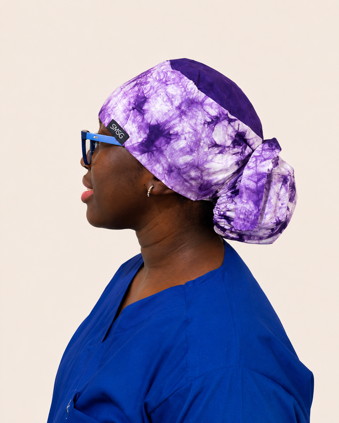 A side-profile portrait of a woman wearing a purple and white tie-dye scrub cap with a small black brand tag that reads 'SMSG'. She is also wearing blue medical scrubs and blue-framed glasses against a solid light beige background.