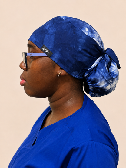 A side-profile portrait of a woman wearing a blue  medical scrub top and a matching blue tie & dye pattern scrub cap. The head covering has a small black brand label that reads 'SMSG'.