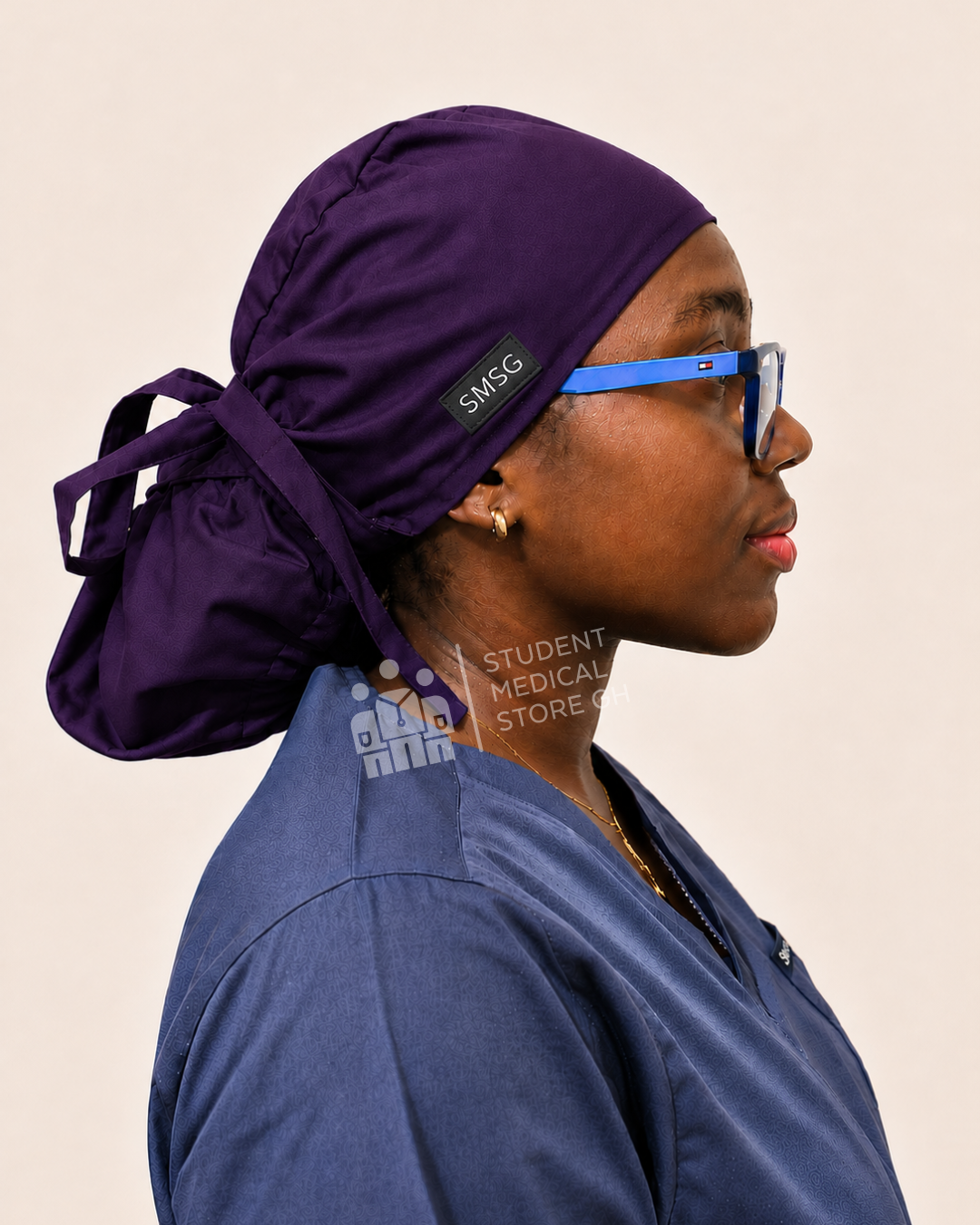 Female nurse wearing grey scrubs  and glasses with a side view of SMSG purple scrub cap.