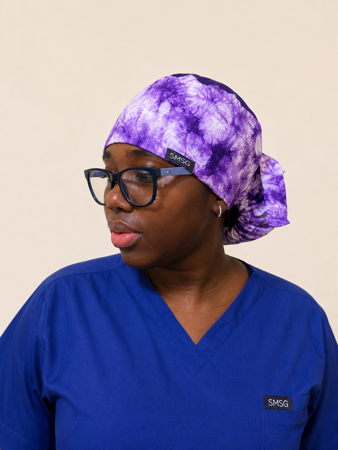 A woman wearing a purple and white tie-dye scrub cap with a small black SMSG logo patch, paired with blue medical scrubs and black-rimmed glasses, set against a solid beige background.